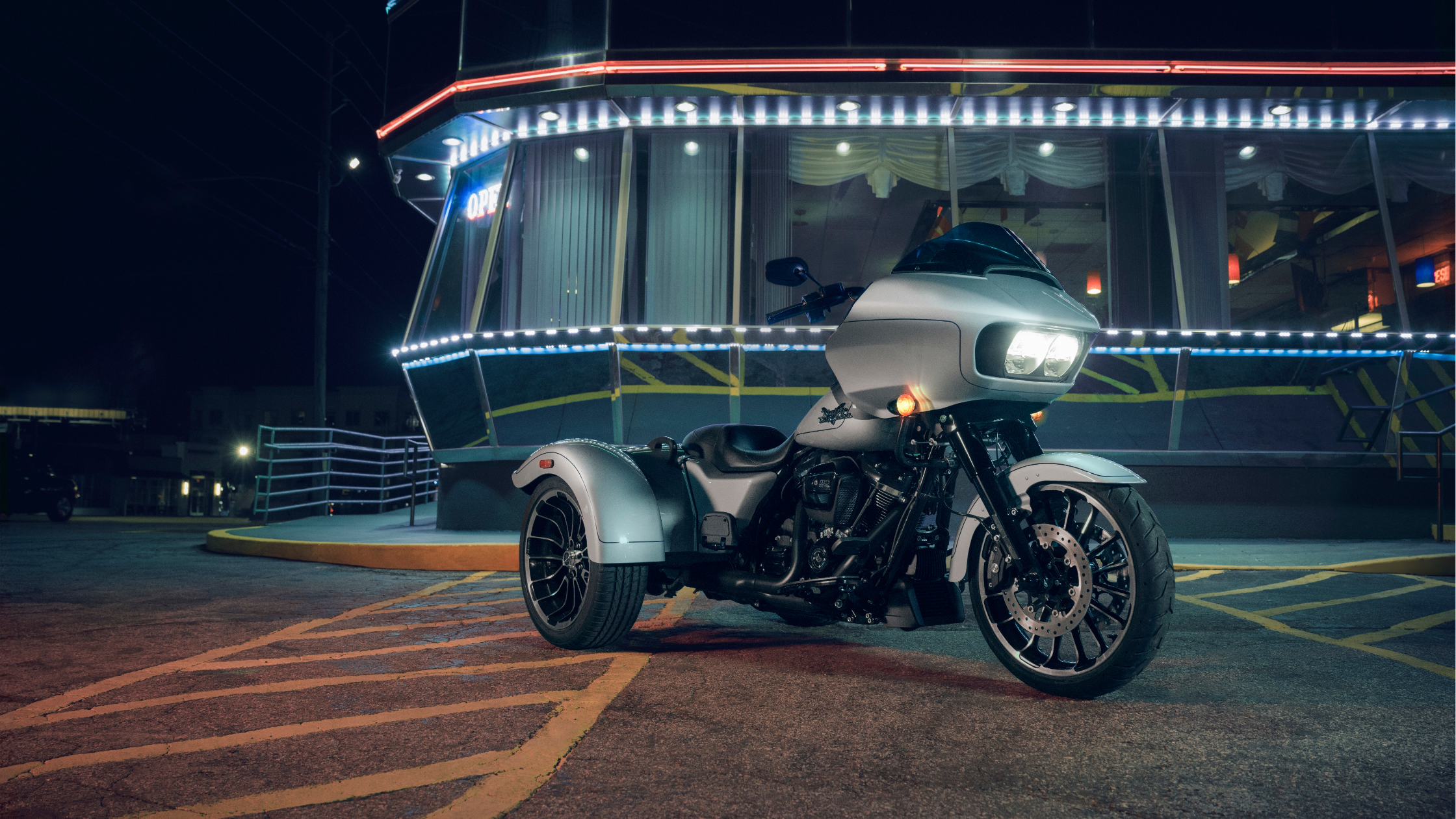 A 2024 Harley-Davidson® trike parked outside a retro diner at night, illuminated by neon lights, sho…